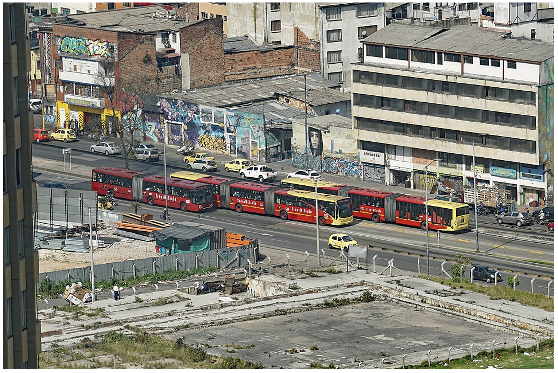 Mehrere rote Gelenkbusse des BRT-Systems TransMilenio fahren auf einer Hauptstraße in Bogotà zwischen Autos und Gebäuden.